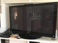 Front view of a large LG flat screen television on a shelf with cables and remote control.