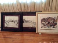 Three framed and matted prints standing on a floor, two monochrome nautical scenes and one colorful still life.