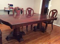 Photo showing wood dining table with carved chairs around it, rectangular shape with two pedestal bases on hardwood floor.