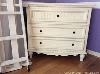 Front view of cream Bassett three drawer dresser with dark hardware and turned legs.