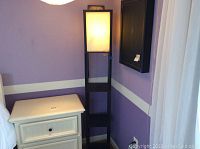 Full view of the three shelf black wood floor lamp with fabric shade, showing its height and shelving design beside a white nightstand in a room with purple walls.