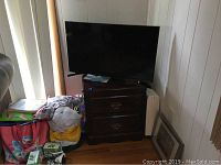Photo showing Samsung TV on top of two drawer dark wood nightstand with some bags and items around