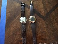 Two vintage wristwatches side by side on wood surface; one gold tone with black face, other silvertone with silver face and leather straps.