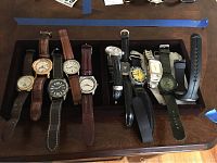 Top view photo of eleven men's watches displayed in a black velvet dresser box on wooden surface, showing various strap and dial styles