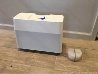 Hard white and light gray plastic carrying case for sewing machine with handle on top, placed on wooden floor alongside a small clear plastic box with purple band.