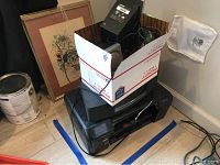 Photo of box and items stacked, includes framed artwork and speakers on top of printer and box