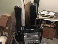 Photo of three black electric appliances: one wheeled heater with visible heating elements, and two tower fans standing behind it, one with manuals on top.