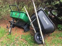 Green John Deere plastic yard cart with hitch and black Truper plastic wheelbarrow side by side outdoors on grass and pine needles.