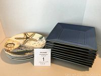 Twelve plates displayed, four round decorative ones featuring various natural and map motifs, stacked beside eight square blue plates.