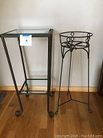 Two tier metal side/accent table with beveled glass shelves and metal plant stand placed side by side on wood floor against white wall.