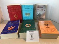 Photo showing six paperback books of the Outlander series by Diana Gabaldon arranged on a white surface, including Outlander, Dragonfly in Amber, Voyager, Drums of Autumn, The Fiery Cross, and A Breath of Snow and Ashes.