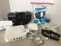 Photo showing Atlantis radio/music player with control buttons, screen, its user manual, multiple white cables including older Apple iPhone chargers and adapters, an Apple Airport Extreme router device with multiple ports, and an Apple Airport Express device.