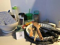 Photo showing a collection of kitchen utensils including wooden spoons, tongs, measuring spoons, electric knife with cord, insulated cups, marble mortar and pestle, and box grater.