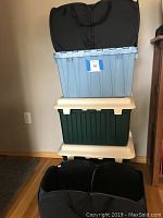 Stack of storage totes including a blue plastic tote on top with lid, two green flip top plastic totes below with lids, and several black fabric trunk organizers at bottom.