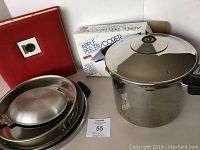 Photo showing stainless steel pressure cooker, apple peeler in box, red recipe notebook, springform pan and frying pans