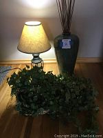 Photo showing lamp lit next to tall glazed ceramic vase with twigs and oblong basket with faux ivy placed on floor.
