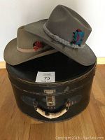 Two Akubra Snowy River hats on top of vintage black leather hatbox, showcasing hat colors and feather decorations.