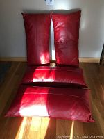 Four rectangular red leather pillows arranged on a wooden floor, sunlight casting shadows