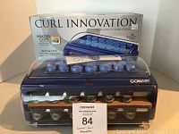 Front view of Conair Curl Innovations curling set in blue heating base with heated clips and clear protective cover, including original box behind it.