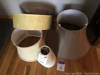 Four various lamp shades clustered on a wooden floor. Large flared beige shade, smaller pale yellow round shade, and two additional shades visible, one with price tag.