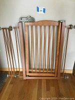 Full view of the pressure-mounted pet gate showing the wooden swinging gate section with vertical slats and metal side rails with mounting hardware attached to the wall.