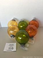 Front view of six glass ornaments in yellow, green, and orange colors arranged on a flat surface with seller's lot card visible