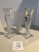 Front view of two clear lead crystal candlesticks with square bases and a frosted triangular glass vase positioned between them