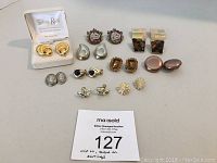 Overview photo showing 11 pairs of earrings arranged in rows including one boxed pair and a variety of clip-on styles in silver, gold tones, amber and cameo designs.