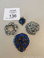 Lot of three pins and one pendant arranged on neutral background. Pins include two clear rhinestone designs and one blue rhinestone cluster. Pendant is heart-shaped blue with faux pearl.