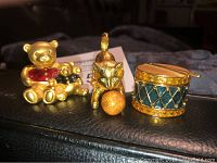 Three gold-tone Estee Lauder perfume compacts: teddy bears, cat, and drum on black surface