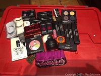 Overhead view of 25 new beauty cosmetic items arranged on a red surface, including boxed MAC products, NARS eyeshadow quad, Clinique acne treatment packet, false eyelashes, and a purple cosmetic pouch.