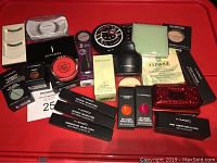 Photo displaying full assortment of the lot on a red tray: makeup powders, lip colors, eyeshadows, pigments, false eyelashes, and boxed skincare products.