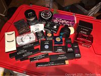 Full view of all cosmetic items arranged on red surface showing boxes, powder jars, compact powders, eye shadows, lip colors, false eyelashes, and more.