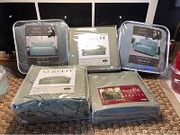Five packaged furniture slipcovers arranged on floor including sofa and loveseat covers by Kathy Ireland Home Essentials and SureFit brand covers in muted green and gray.