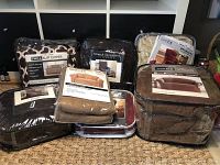 Seven packaged furniture slip covers arranged on woven mat floor in front of shelving. Various brands and types including twill, stretch sensations, homestyle, and sanctuary as seen on packaging.