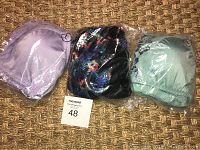 Three Rhonda Shear bras in original packaging laid on textured surface. Two solid colors (lavender and mint green) and one floral pattern near center.