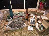 Photo showing assorted home decor including large metal leaf tray, metal trinket holders, blue glass vase, ceramic cactus piece, wooden figurine, bathroom accessories, gold-toned photo frame, and boxed items.