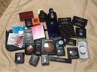 Photo showing 25 makeup and beauty products including powders, eyeshadows, lip colors, blushes, samples and a makeup pouch spread on a beige surface