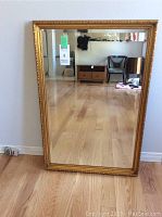 Front view of the large rectangular mirror with ornate gold frame standing on wooden floor, showing full mirror and some room reflection.