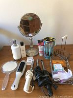 Photo showing curling irons, makeup mirror, lotion dispensers, soaps, sleep mask, and assorted toiletry accessories on table.