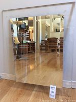 Front view of the rectangular bevelled mirror with reflective surface and acrylic frame resting against the wall on a wooden floor.