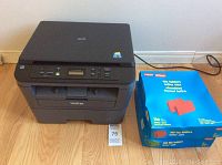 Brother DCP L2520DW printer with power cord, shows front top and control panel. Two unopened boxes of red file folders to the right.