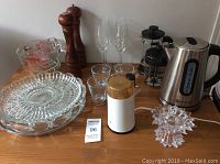 Full view of all kitchen items on wood table: coffee grinder, electric kettle, Bodum French presses, glassware, measuring cups, wooden pepper grinder, cake stand and candle holder.