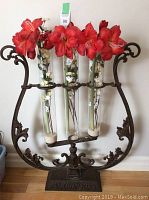 Full front view showing ornate cast iron frame shaped like a lyre holding three clear glass tubes with artificial flowers inside.
