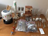 Overall view of the lot on a wooden table showing the fondue set, hand mixer, cutlery in plastic bags, salt and pepper shakers, nut chopper box, and glass tumblers.