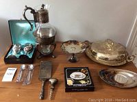 Display of all silver plated items on a wooden table showing variety of serving pieces, flatware, goblets, and pitcher with stand.