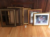 Seven picture frames in total, showing two gold framed prints and several empty frames leaning against wooden furniture on hardwood floor.
