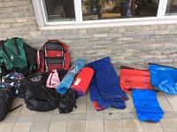 Full view of camping gear including backpacks, sleeping mats, and dry sacs laid out on tile floor under window