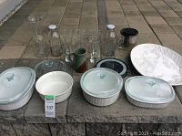 Full lot displayed on outdoor stone surface showing Corningware dishes, glass bottles, loose leaf teapot, ceramic mug, and serving plate with chip.