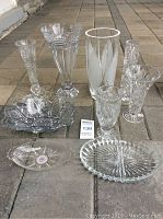 Group shot of nine assorted cut crystal and glassware pieces arranged on patio floor, including vases and dishes with intricate cut patterns.